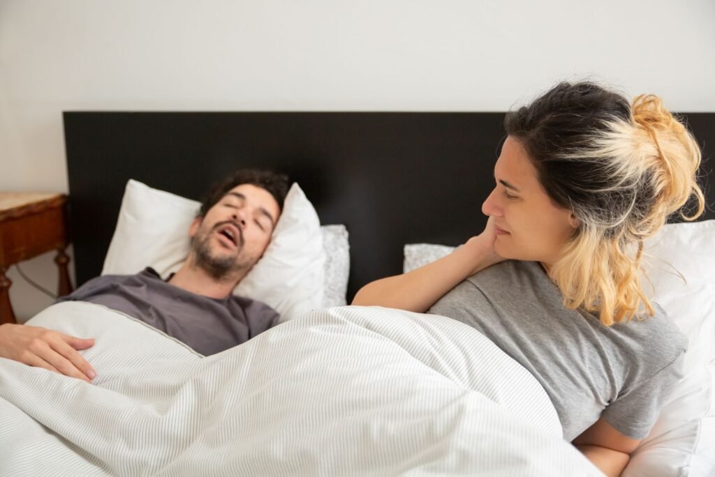 Man snoring in bed while his partner lies awake, a common sign of sleep apnea